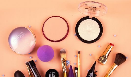 Makeup kit with cosmetic products including foundation, brushes, lipstick, mascara, powder, and beauty blender on a peach background